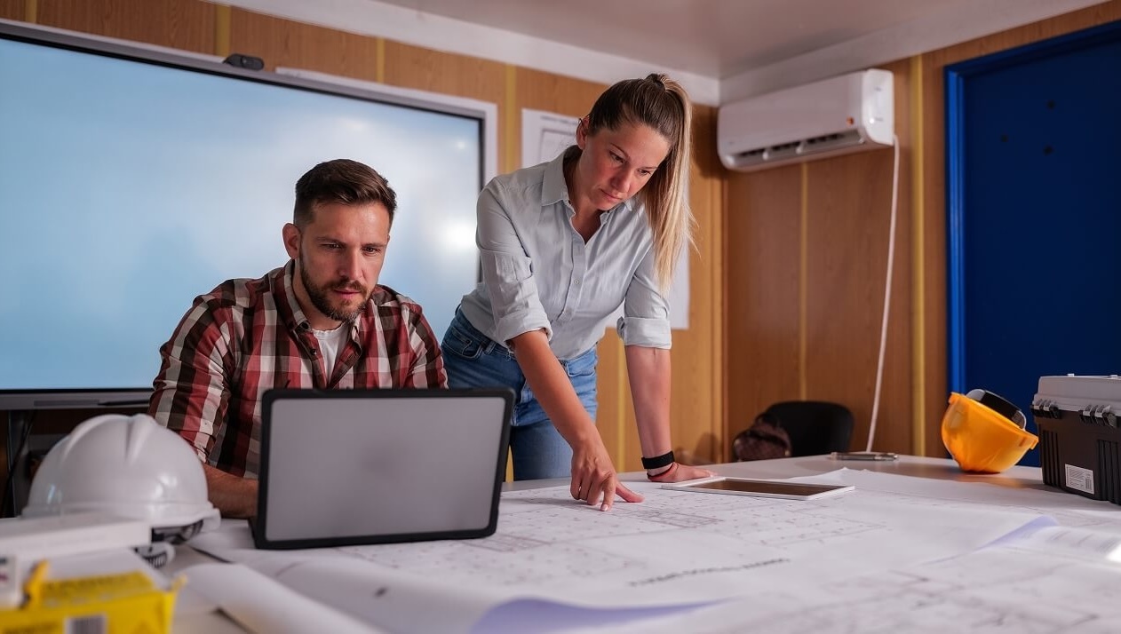 Two construction professionals reviewing blueprints and a laptop in a site office with hard hats and a toolbox on the table.