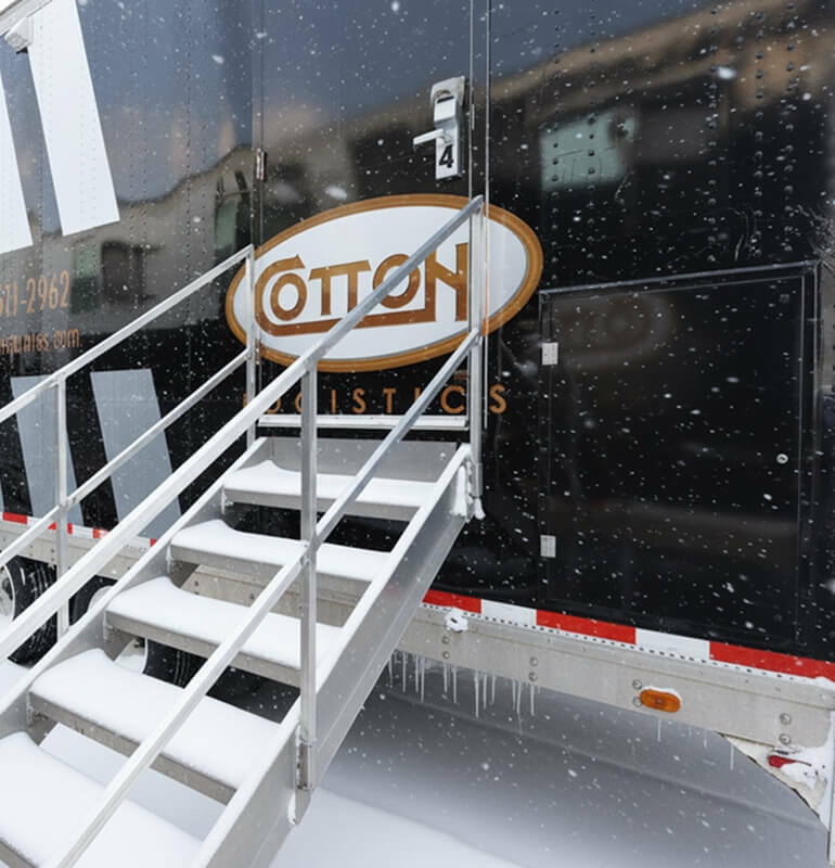 A close-up of snow-covered metal stairs leading up to the door of a black Cotton Logistics trailer during a snowstorm.
